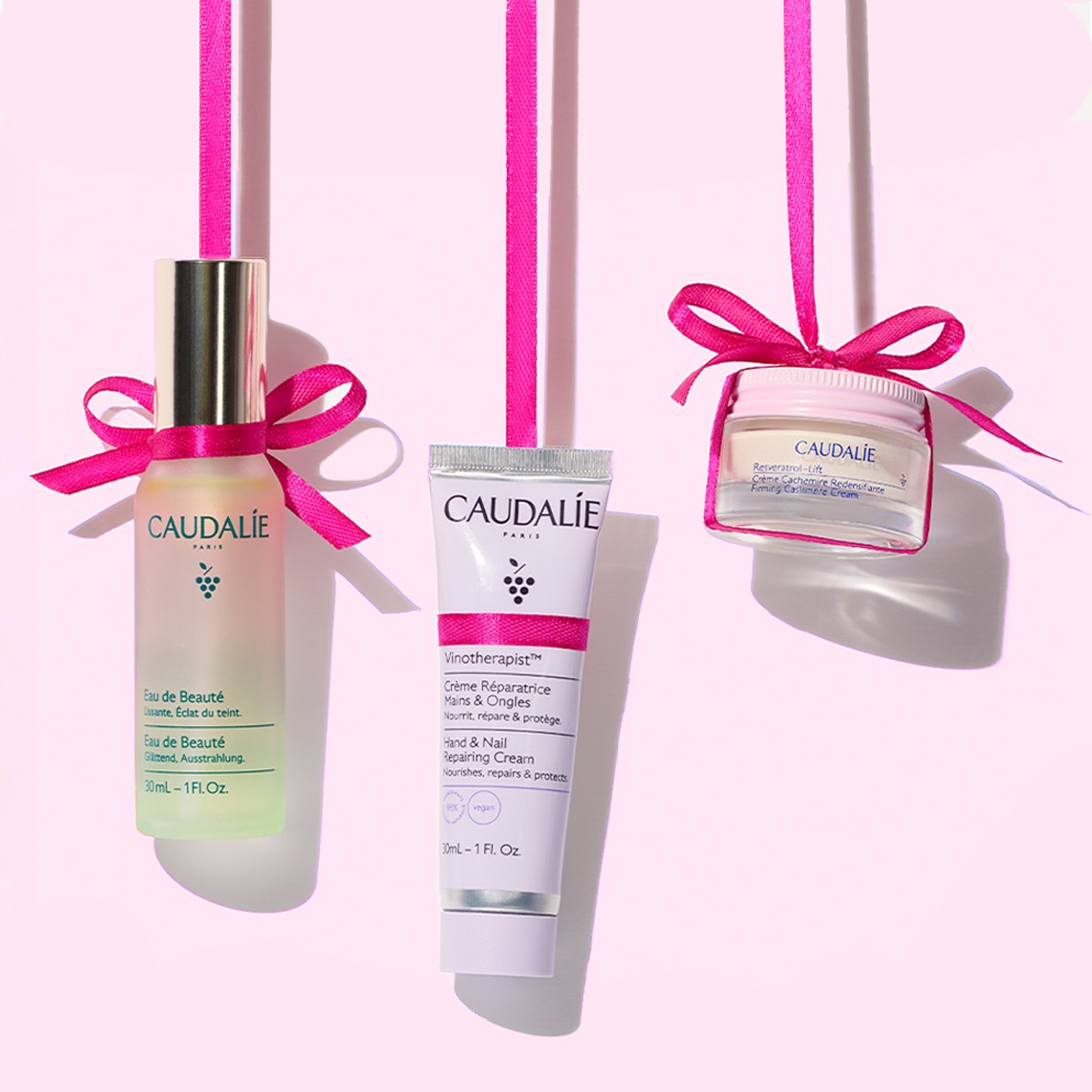 Three Caudalie skincare products with pink ribbons hang against a light pink background.