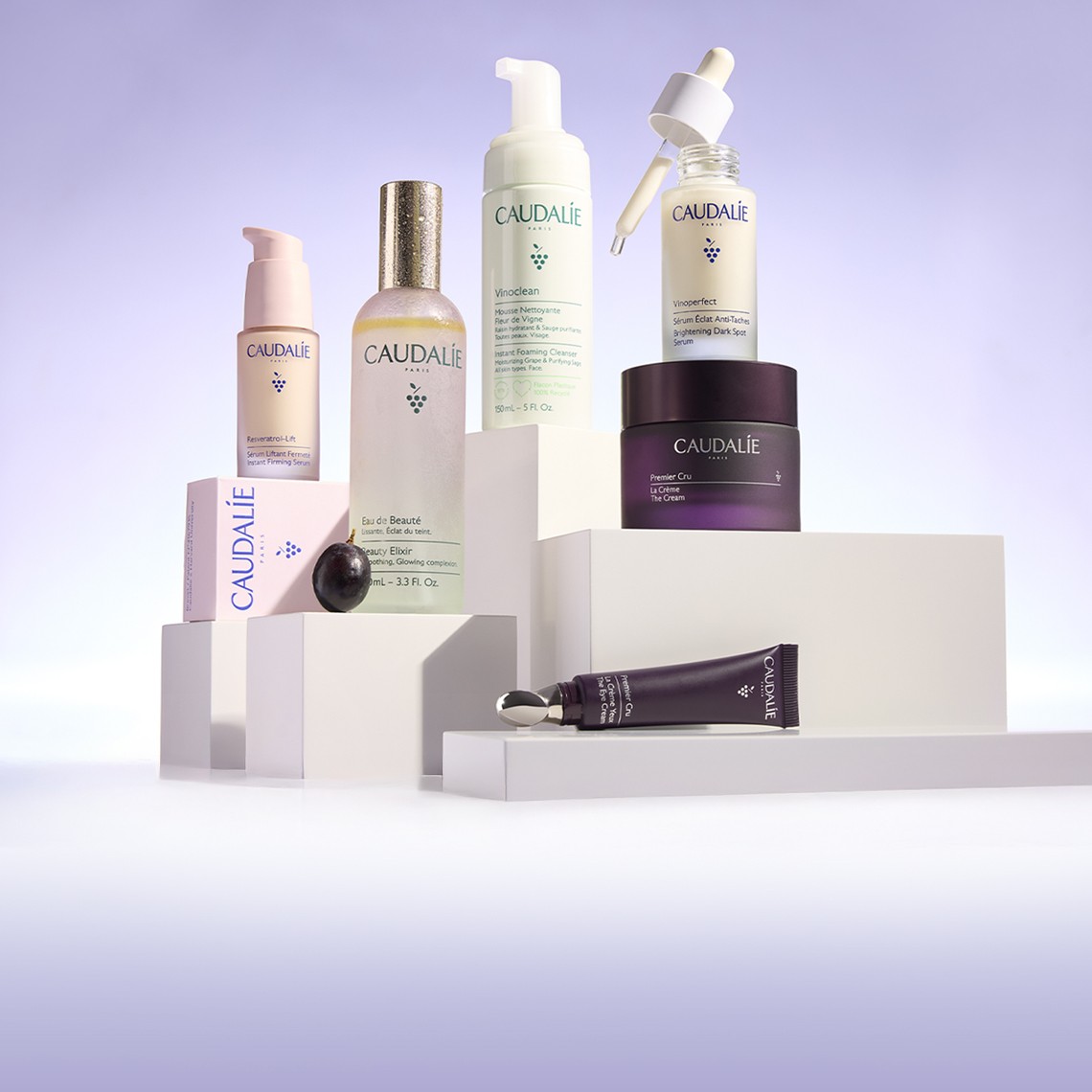 A collection of Caudalie skincare products, including bottles and tubes, displayed on white platforms against a purple gradient background.