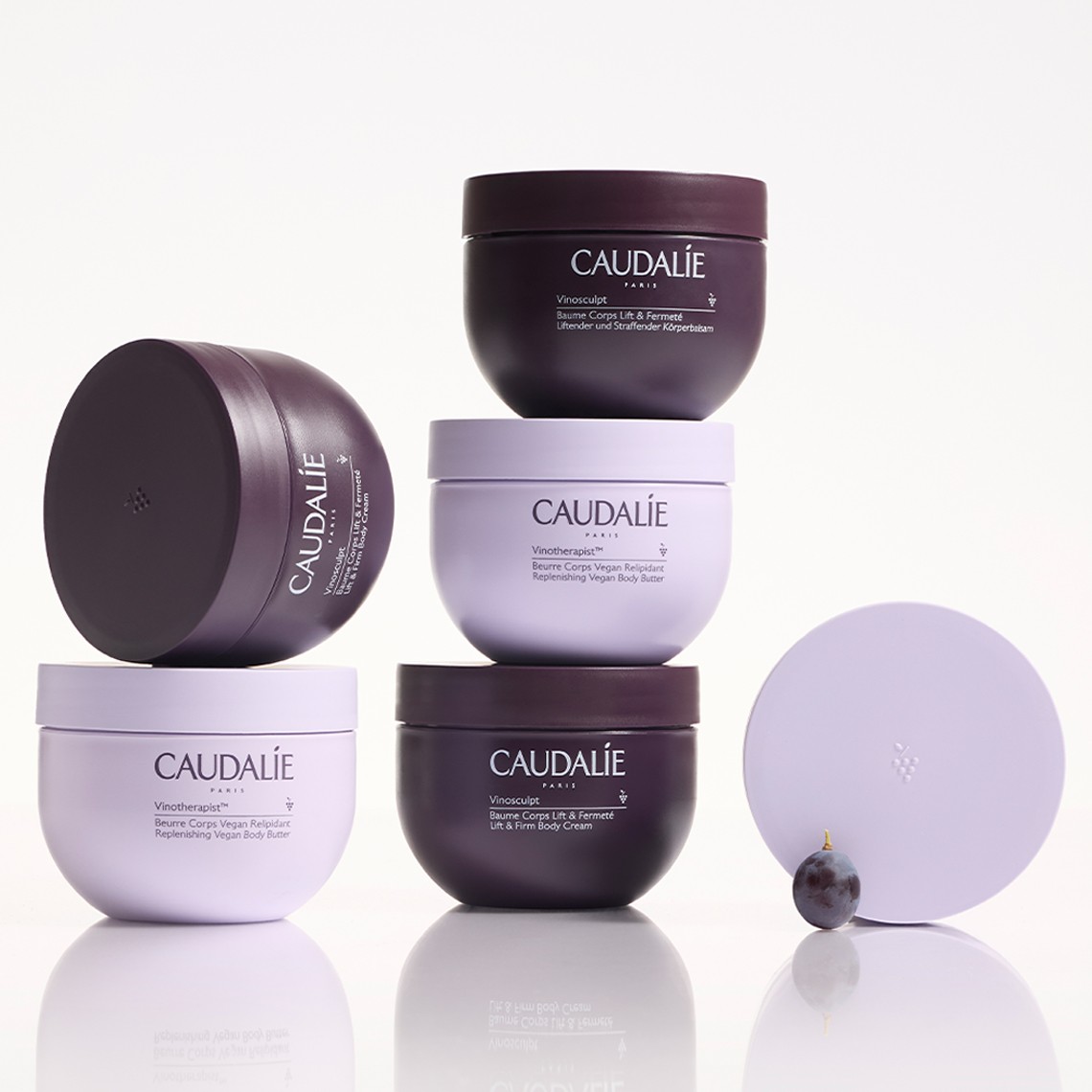 Stacked Caudalie creams in purple and white containers on a reflective surface, with a small dark grape beside them.