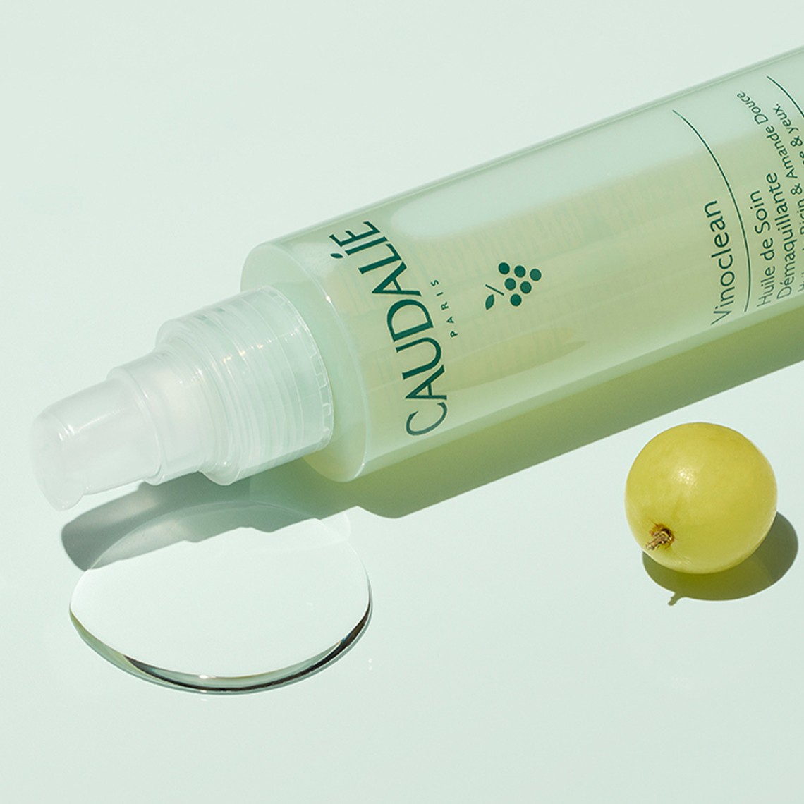 A spray bottle labeled "Caudalie" lies on its side next to a small yellow-green fruit and a clear liquid droplet on a light green background.