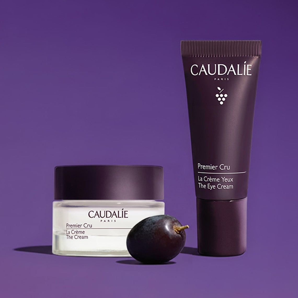Two Caudalie skincare products, a jar and a tube from the Premier Cru line, are displayed on a purple surface with a grape in front.