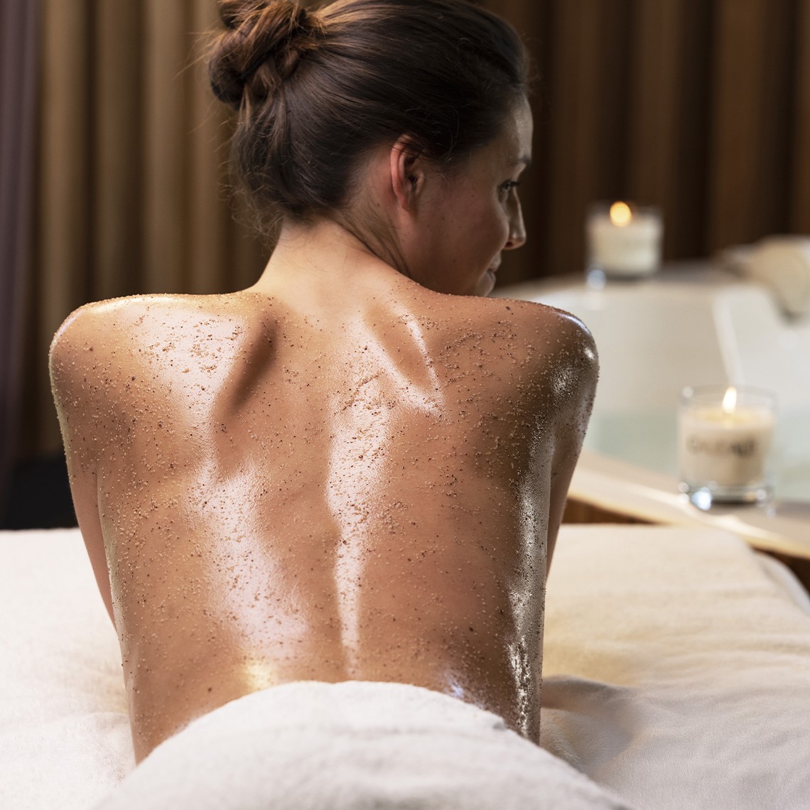Person face down on spa table with exfoliating scrub on bare back, towel at hips and lit candles nearby.