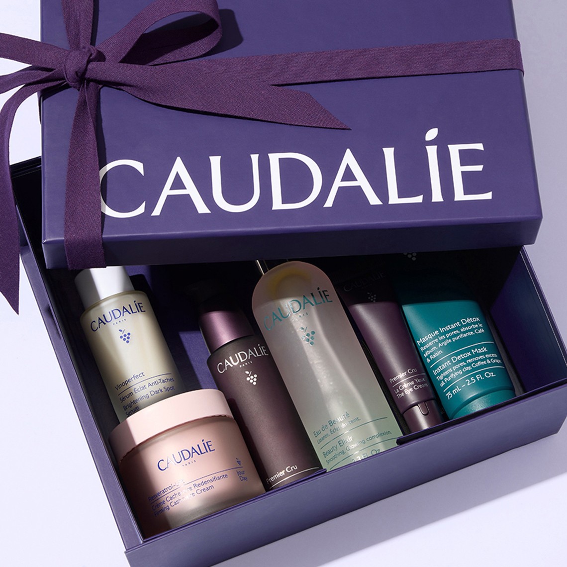 Purple gift box with "Caudalie" branding, filled with skincare products, tied with a matching ribbon, on a light purple background.