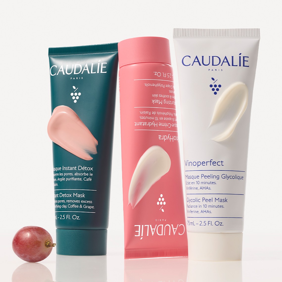 Three Caudalie product tubes stand upright, featuring pink and white creams, with a single red grape nearby on a white background.