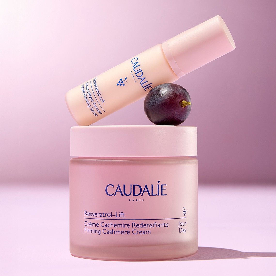 Caudalie Resveratrol-Lift cream and serum on a pink background, with a grape beside them.