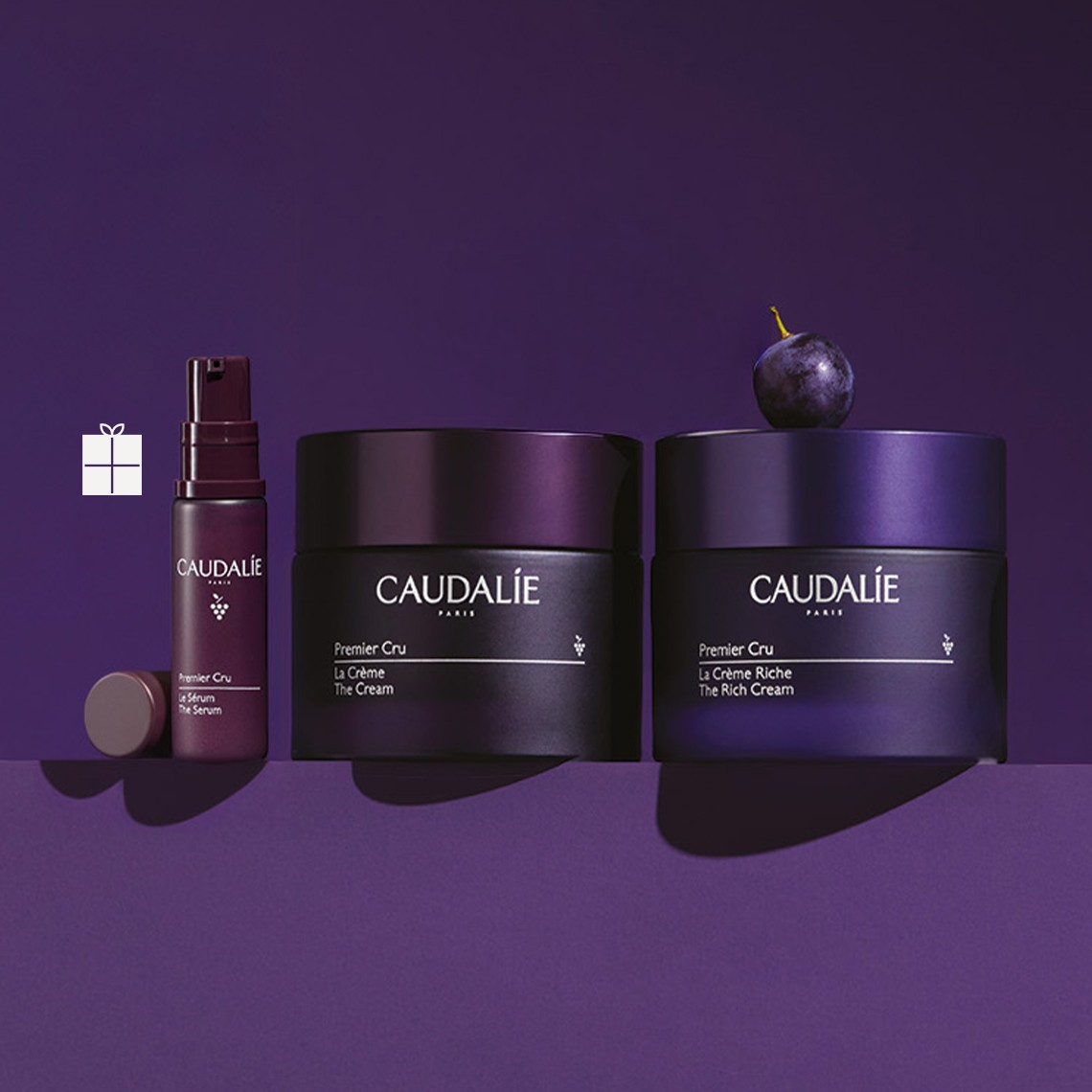 Purple skincare products labeled "Caudalie" with two cream jars, a serum bottle, and a grape, set against a deep purple background.