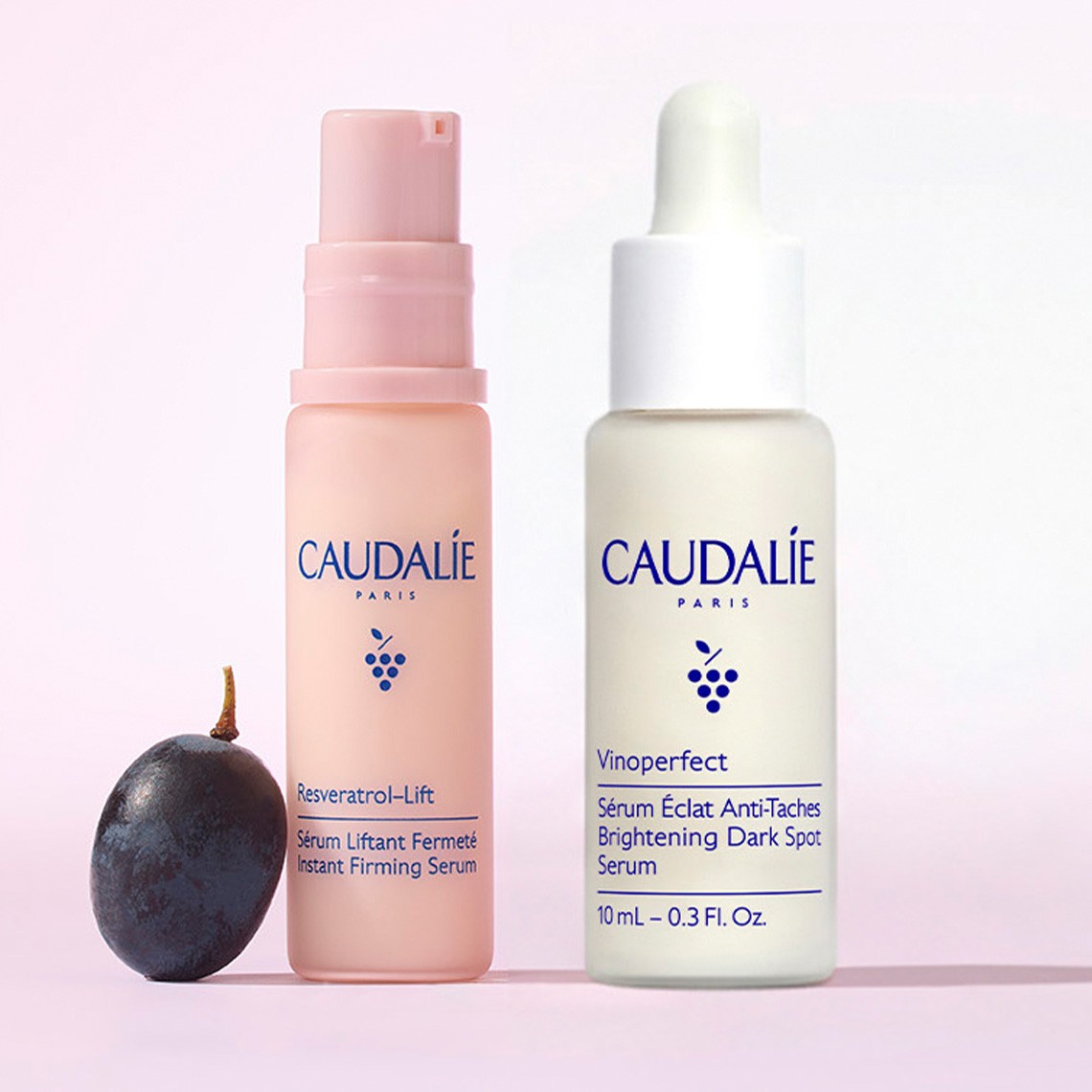 Two Caudalie skincare bottles, one pink and one white, with a grape on a pink background.