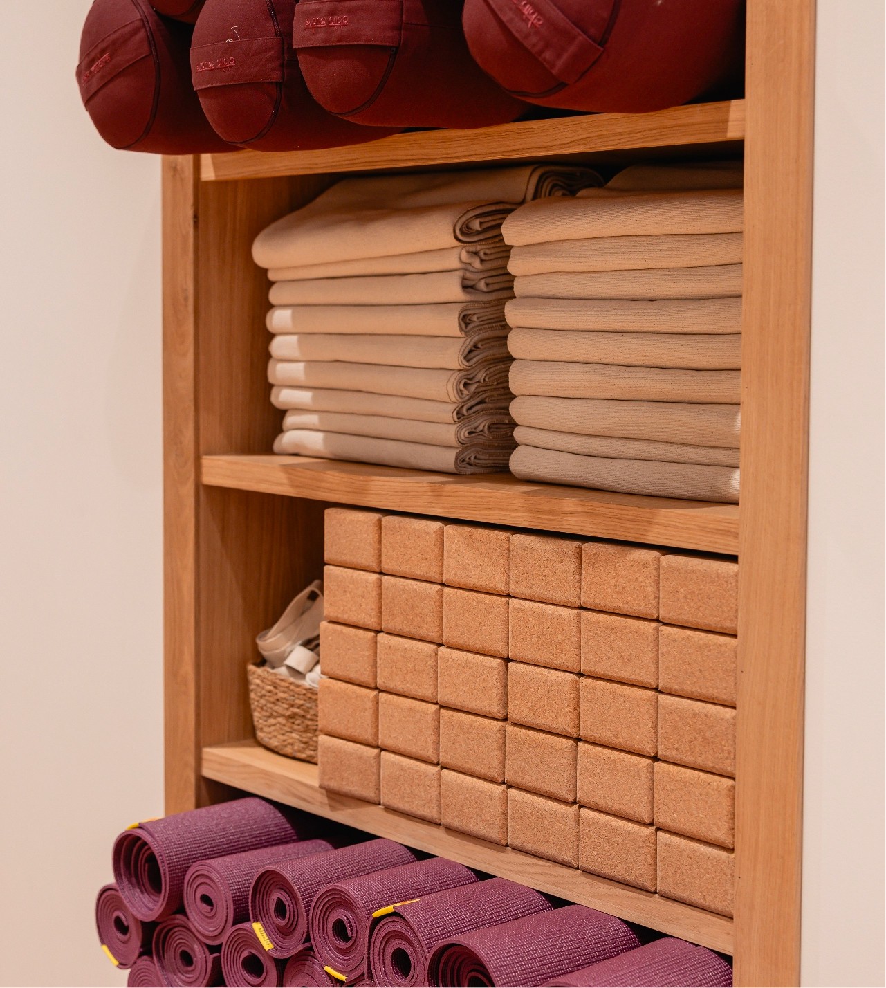 Wooden shelf with yoga mats, towels, cork blocks, and baskets neatly organized. Mats are purple, towels beige, blocks in a grid pattern.