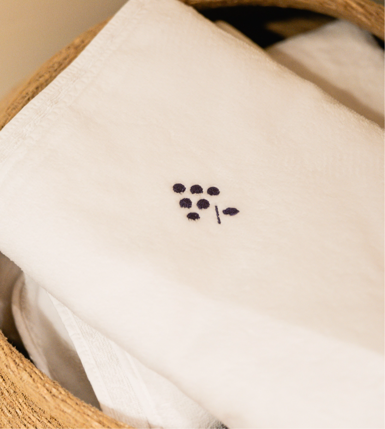 White towel with a small blue embroidered grape design folded in a wicker basket.