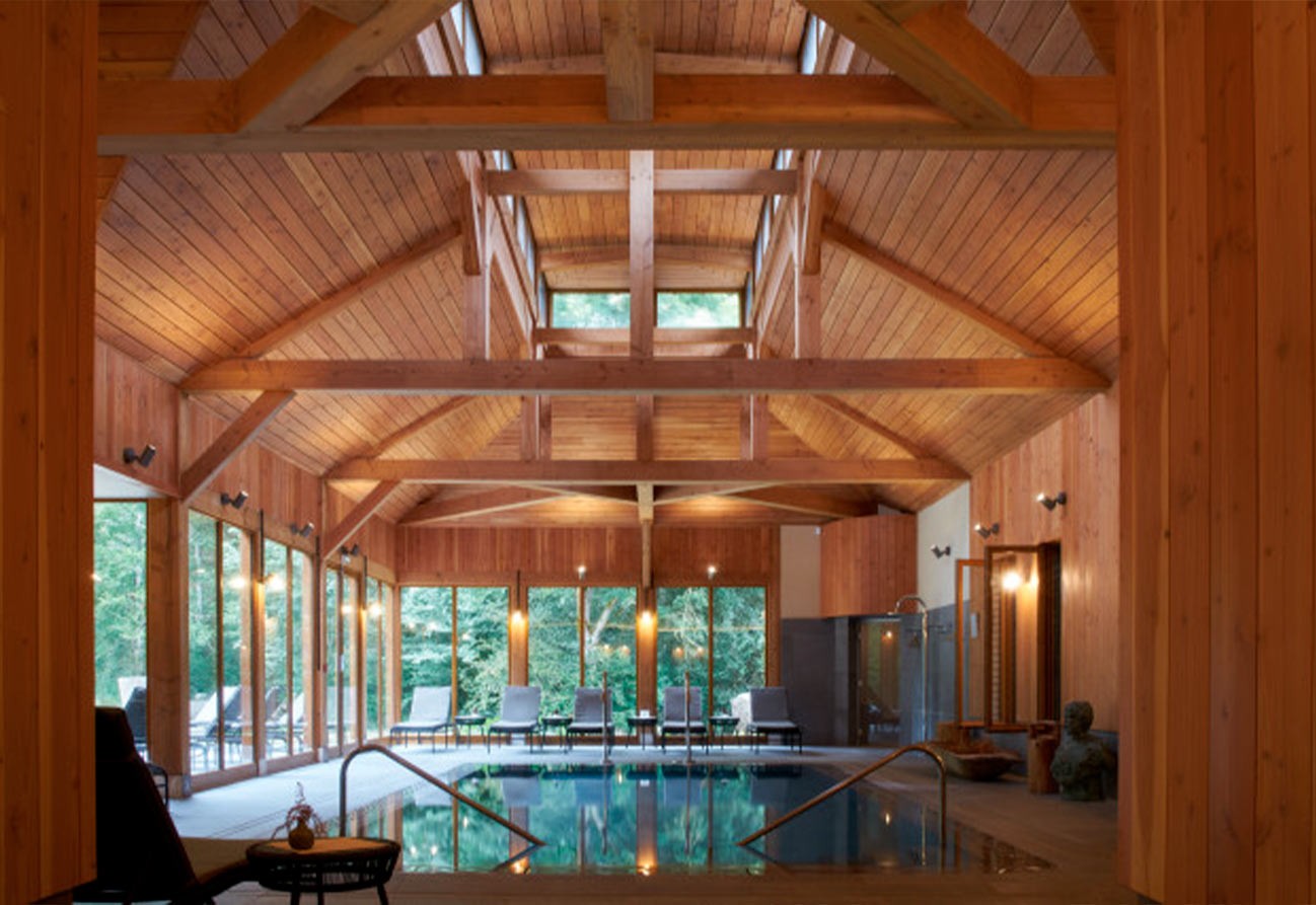 Indoor wooden-beamed pool room with high vaulted ceiling, large windows, lounge chairs and a reflecting pool overlooking forest views.