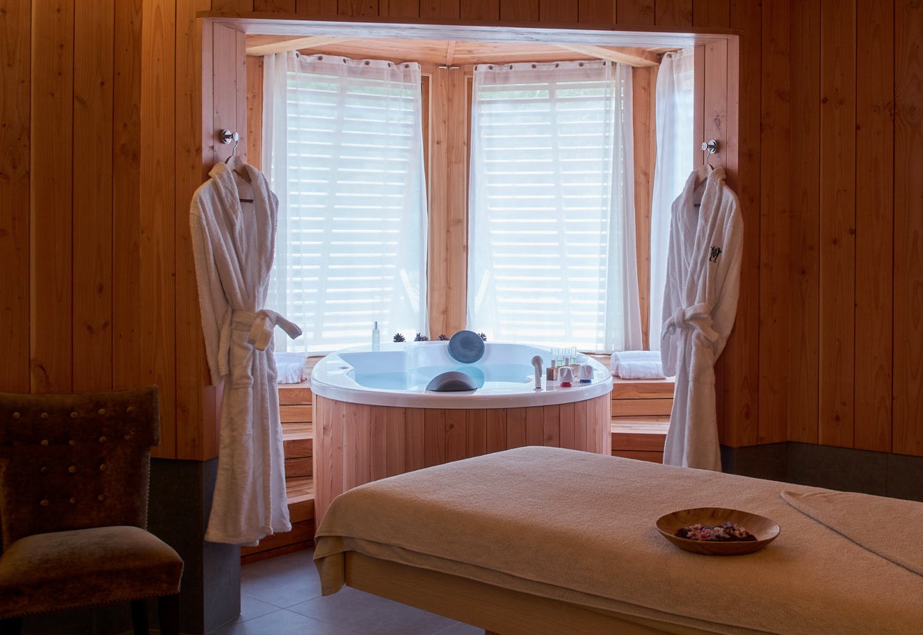 Cozy wooden spa room with round jacuzzi under a bay window, two white robes hanging and a massage table with a wooden bowl.