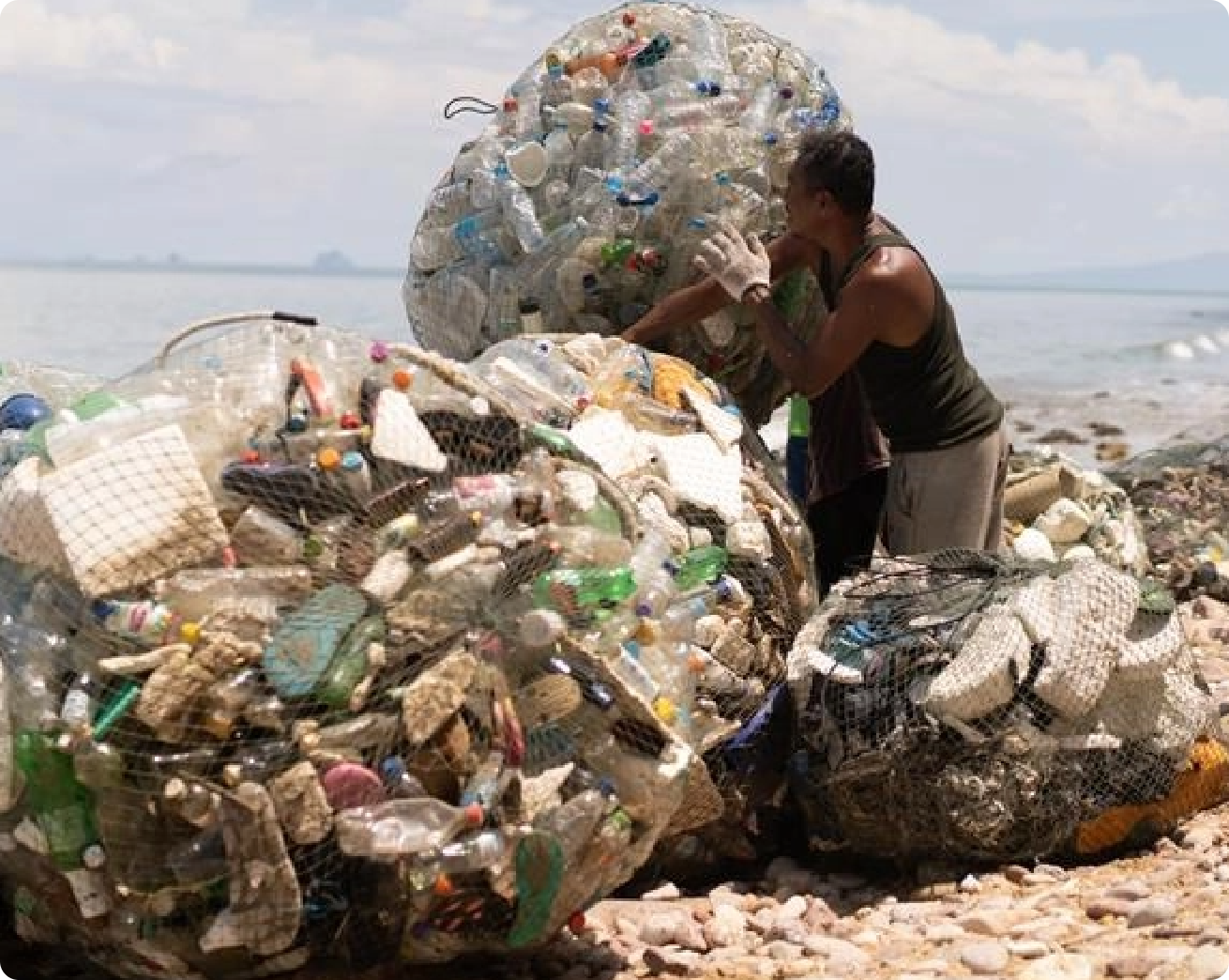 A person on a rocky beach sorts large, enclosed bundles of plastic waste under a cloudy sky.