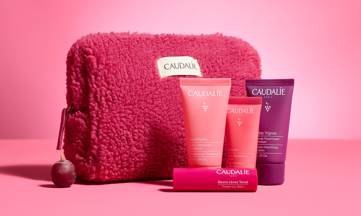 Pink textured pouch with four Caudalie skincare products, including creams and a lip balm, set against a pink background.