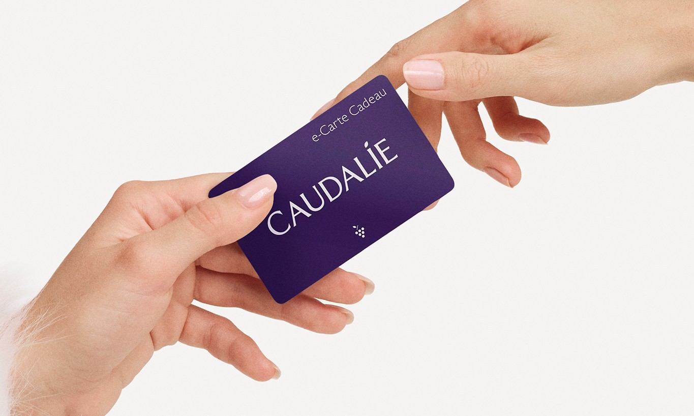 Two hands exchanging a purple gift card with "CAUDALÍE" printed on it, against a white background.