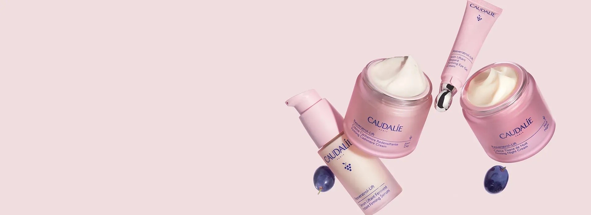 Caudalie skincare products displayed on pink background including cream jars, serum bottle, and eye care tube with blueberries as styling elements