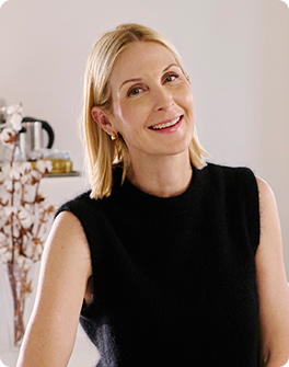 Smiling woman with short blonde hair in a sleeveless black top, standing indoors with a minimalistic background.
