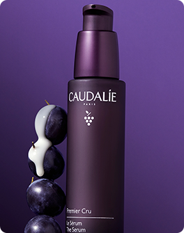 A dark purple bottle of Caudalie Premier Cru Serum against a purple background, accompanied by stacked grapes with cream drizzling over them.