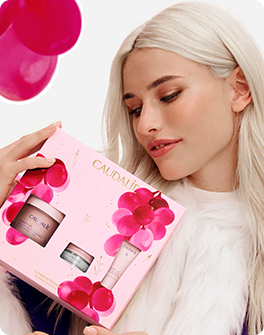 Woman with long blonde hair holding a pink Caudalie gift set, featuring skincare products, adorned with grape illustrations.
