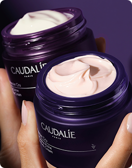 Two open purple jars of Caudalie face cream, one white and one pink, held by a hand against a dark purple background.