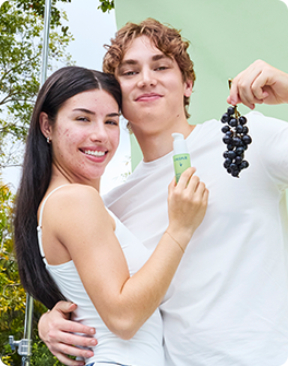 A smiling couple holds skincare products and a bunch of grapes outdoors, standing against a green backdrop.