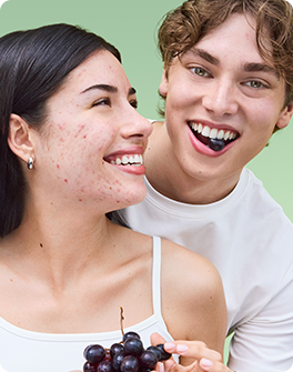 A smiling woman with acne holds grapes, while a man beside her eats one, both against a light green background.