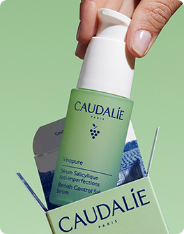 A hand holding a green bottle of Caudalie Vinopure Blemish Control Serum, partially removed from its packaging, against a green background.