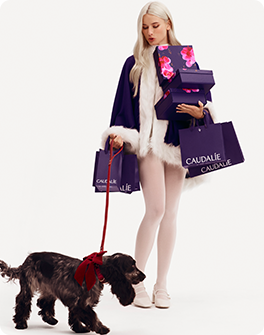 Woman in a purple coat holds Caudalie gift bags and boxes, with a dog on a red leash.