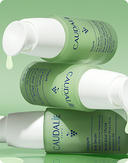 Three stacked green bottles of Caudalie skincare product with pumps, set against a green background. Liquid drips from the top bottle.