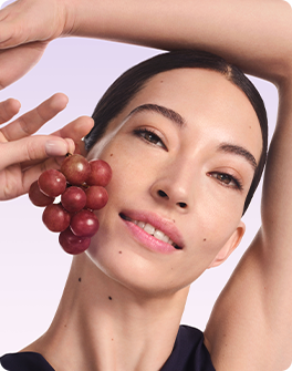 Woman with glowing skin holding red grapes near her face against a light purple background.