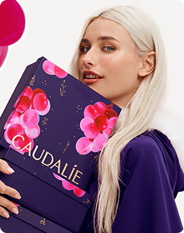 A person with long blonde hair holds a purple Caudalie gift box decorated with pink grapes, wearing a purple outfit.
