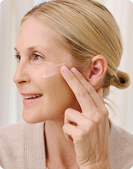 Woman applying moisturizer to her cheek, smiling, with her hair tied back, wearing a beige sweater, in a softly lit environment.