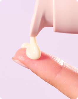A hand dispensing a creamy beige skincare product onto a person's arm against a pink background.