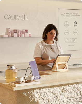 A woman at a Caudalie store counter using a tablet, with skincare products displayed on a shelf and a teapot with cups nearby.