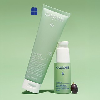 Caudalie skincare products displayed on a green background, including a cleansing gel tube, facial serum bottle, and a small gift box with blue ribbon.