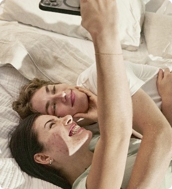 A smiling couple lying on a bed, taking a selfie together with a smartphone.