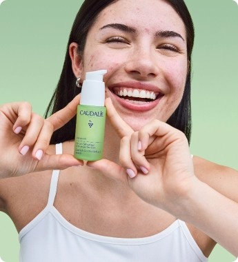 Smiling woman holding a Caudalie skincare bottle against a green background, wearing a white tank top.