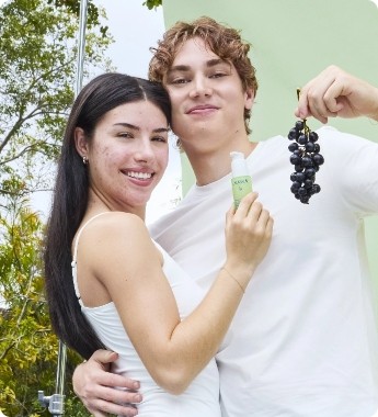 A smiling couple outdoors, the woman holds a skincare product, and the man holds a bunch of grapes. Both are wearing white tops.