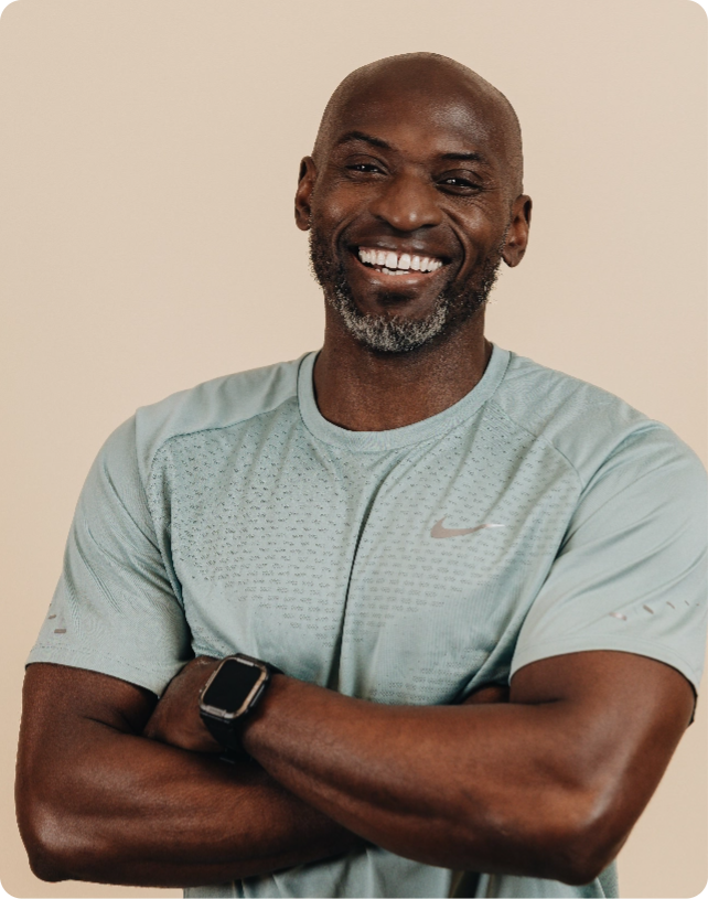 Smiling man with a beard and short hair, wearing a light green athletic shirt and smartwatch, arms crossed against a beige background.