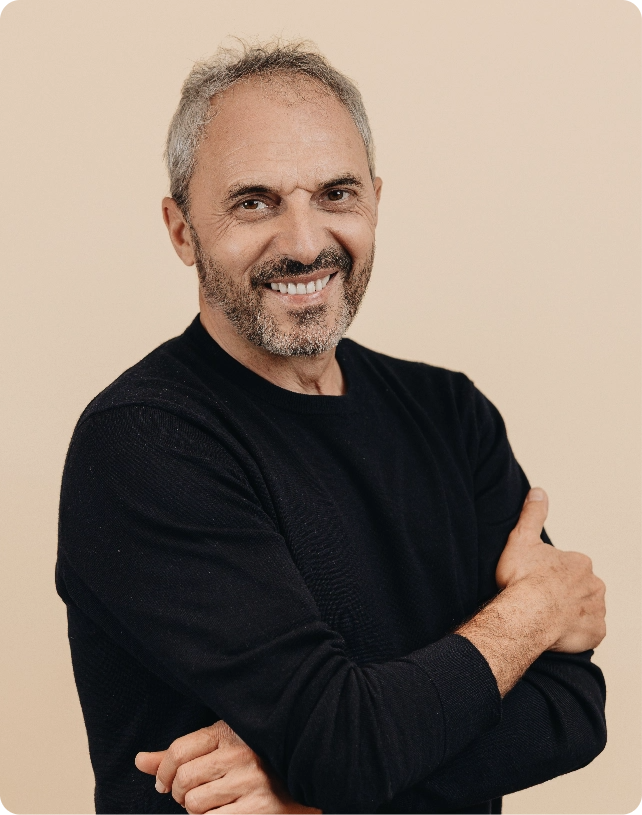 Smiling man with gray hair and beard wearing a black sweater, arms crossed, against a beige background.