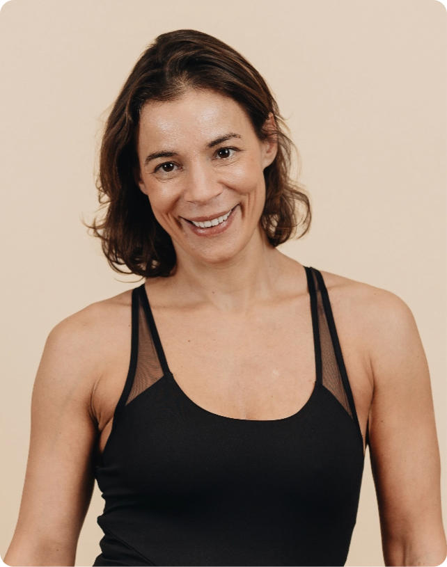 Smiling woman in a black tank top stands against a beige background, looking directly at the camera.