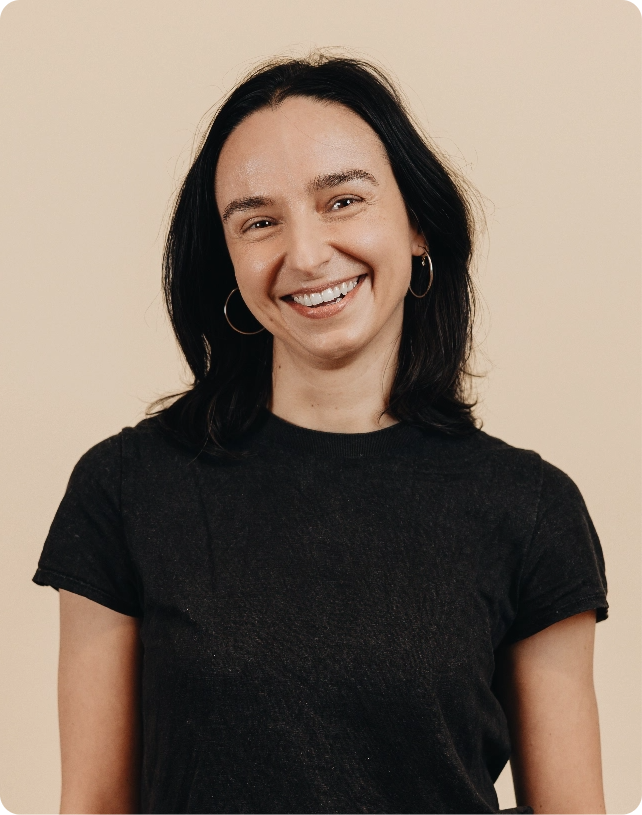 A smiling person with long dark hair, wearing hoop earrings and a black T-shirt, poses against a beige background.
