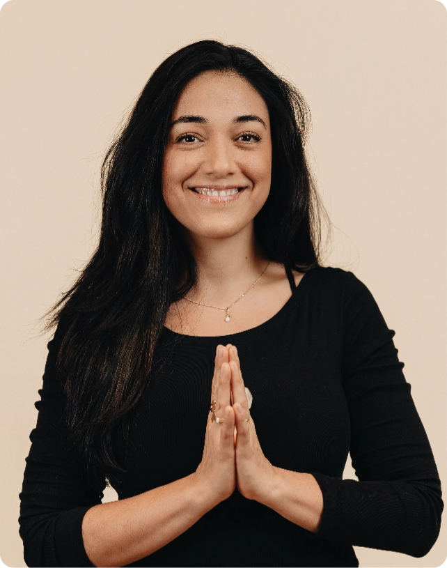 Woman in a black top smiling, with hands in a prayer position against a beige background.