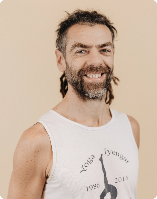 A man with a beard and dreadlocks smiles, wearing a white tank top with "Yoga Iyengar" printed on it, against a beige background.