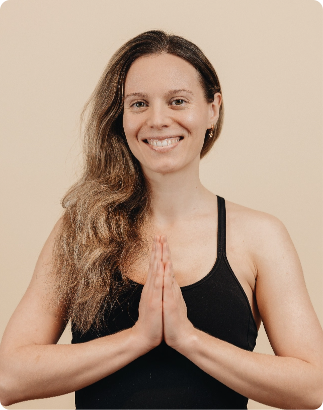 Smiling woman in a black tank top with long hair, holding hands in prayer position against a beige background.