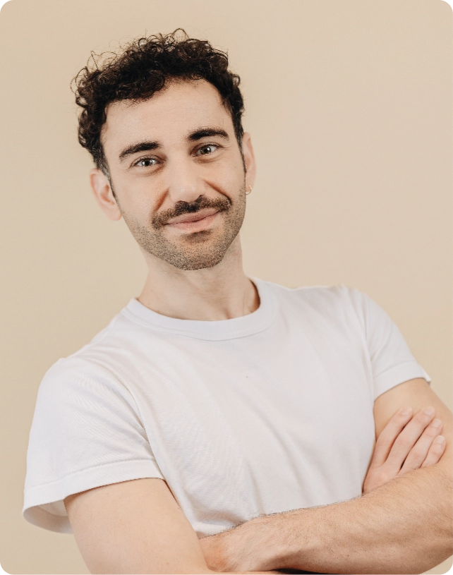 Person with curly hair and a mustache smiles, wearing a white t-shirt with arms crossed, posed against a beige background.