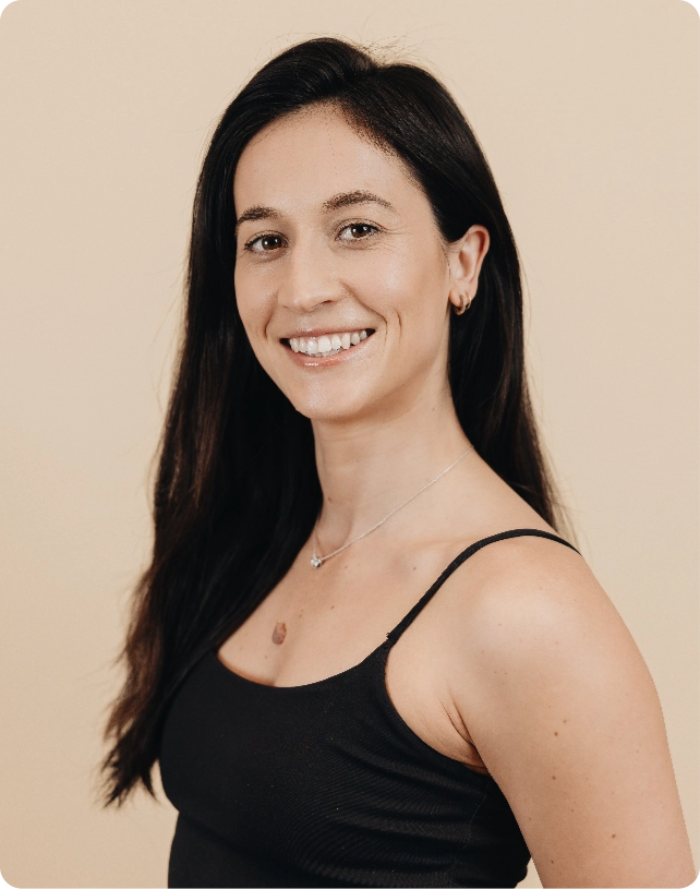 Woman with long dark hair smiling, wearing a black sleeveless top and a necklace, set against a light beige background.