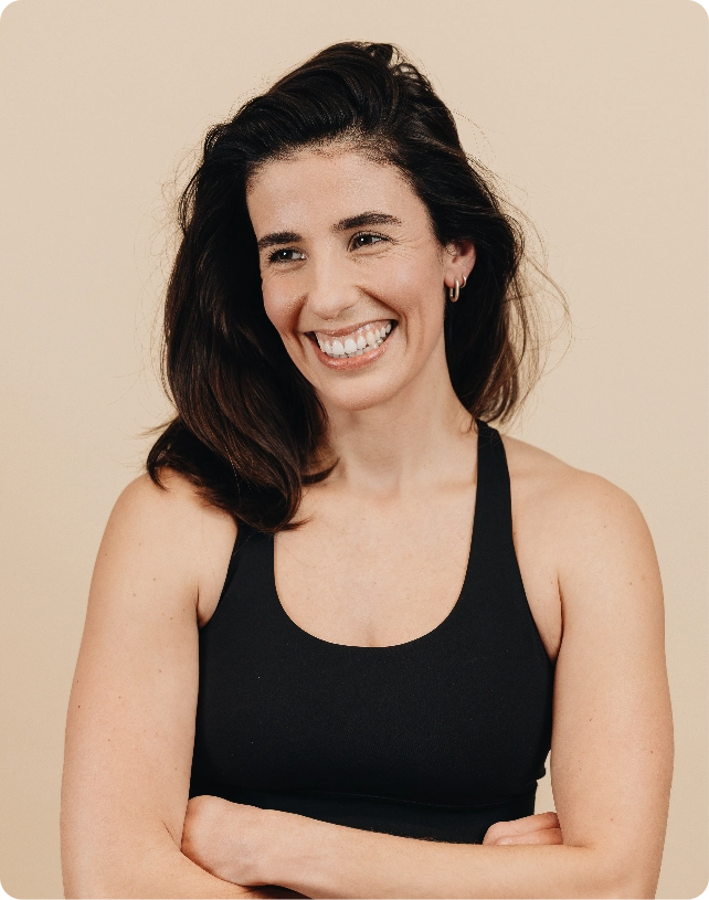 Woman with dark hair smiles warmly, wearing a black tank top against a beige background. Arms are folded casually.