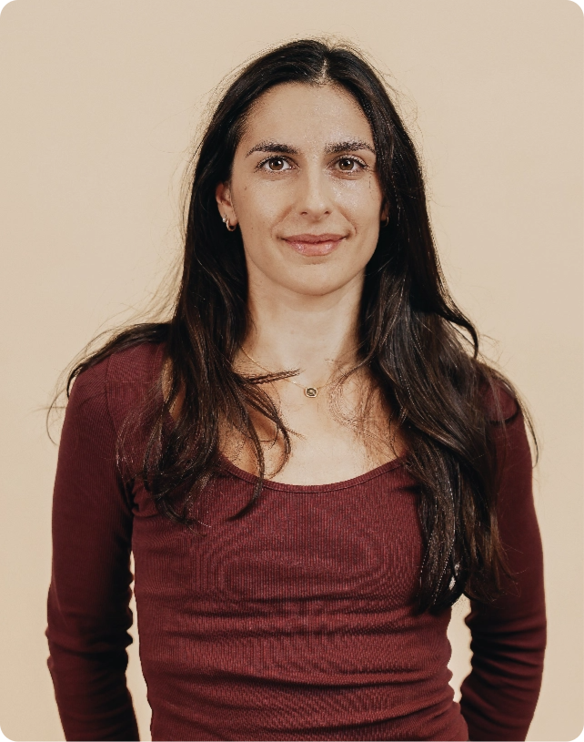 Woman with long dark hair wearing a maroon top, standing against a beige background, smiling softly.