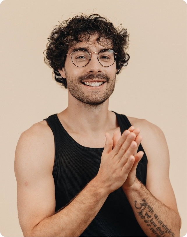 Smiling man with curly hair and glasses, wearing a black tank top. He has a tattoo on his forearm and is clapping his hands together.