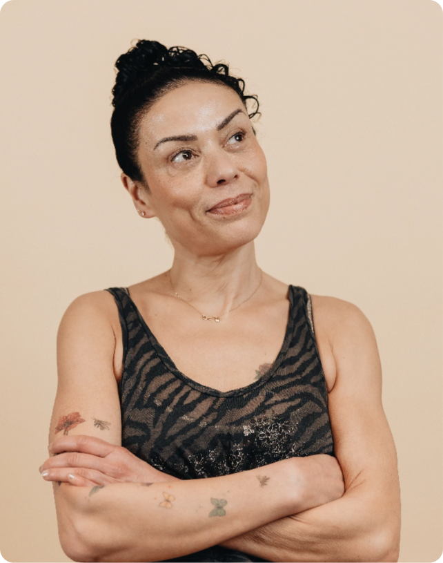 Woman with tattoos on arms, wearing a patterned sleeveless top, looking to the side with a neutral expression against a light beige background.